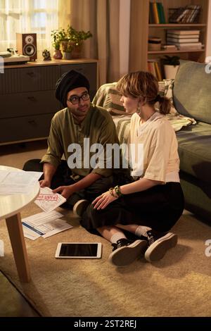 Jeune homme et jeune femme s'engageant dans la conversation tout en examinant des documents à côté d'une tablette sur le sol dans un salon informel avec des sièges confortables et des étagères Banque D'Images