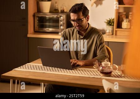 Homme dans la cuisine travaillant sur ordinateur portable, entouré d'un éclairage chaleureux et d'un décor confortable, indiquant une atmosphère détendue mais concentrée dans le cadre du bureau à domicile Banque D'Images