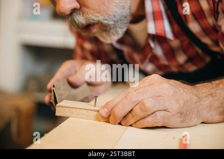 Un menuisier senior en chemise à carreaux rouge sable soigneusement un morceau de bois dans son atelier. Les outils pendent Banque D'Images