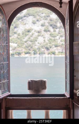 Une fenêtre arquée ouverte avec verre décoratif offre une vue tranquille sur un lac calme entouré de collines verdoyantes, évoquant un sentiment de paix et de détente Banque D'Images