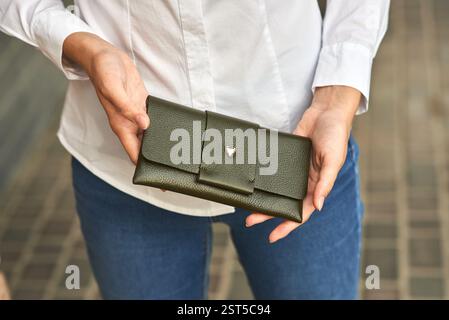 Portefeuille élégant en cuir vert tenu par une femme en chemise blanche et Jean bleu, concept de scène de rue extérieure Banque D'Images