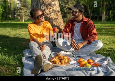Jeune couple noir assis sur une couverture de pique-nique, manger et discuter dans le parc. Date détendue dans la nature, escapade urbaine Banque D'Images