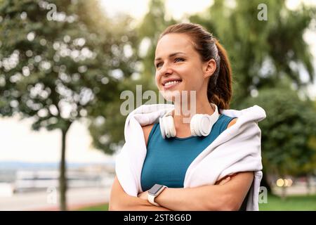 Portrait d'une jeune femme adulte en forme avec des écouteurs autour du cou, marchant à l'extérieur et souriant Banque D'Images