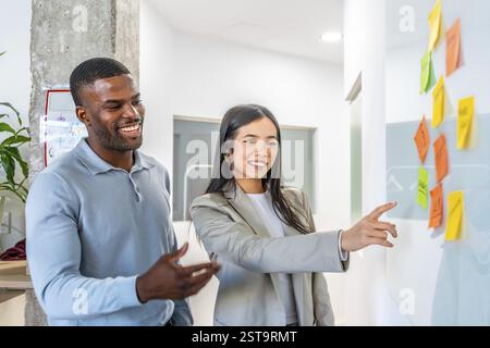 Hommes d'affaires souriants analysant le plan de projet à l'aide de notes autocollantes colorées sur tableau blanc dans un bureau de coworking moderne Banque D'Images