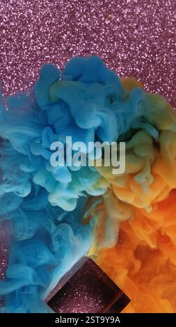 Fumée multicolore. Éclatement de peinture. La brume de flou bleu orange se propage sur fond abstrait rose de paillettes. Cube de verre brillant dans l'eau. Banque D'Images