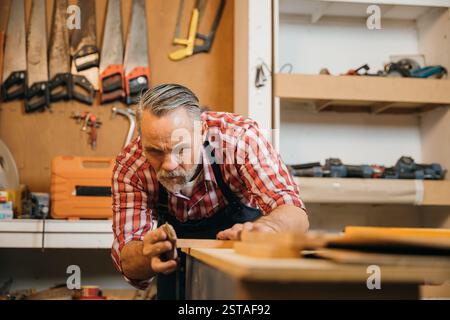Un menuisier senior en chemise à carreaux rouge sable soigneusement un morceau de bois dans son atelier. Les outils pendent Banque D'Images