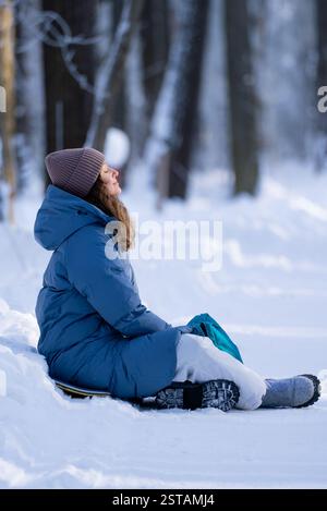 Jeune femme portant des vêtements chauds d'hiver est assise sur un snowboard dans une forêt enneigée, prenant une profonde respiration et profitant du paysage hivernal paisible Banque D'Images