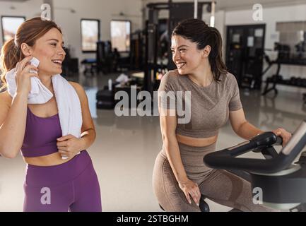 Deux femmes bavardent joyeusement après s'être entraînées dans un gymnase bien équipé, l'une sur un vélo d'appartement et l'autre essuyant la sueur de son front Banque D'Images