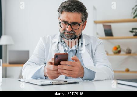 Le médecin masculin est assis avec le téléphone dans les mains. Un travailleur médical professionnel en manteau blanc est dans le bureau. Banque D'Images