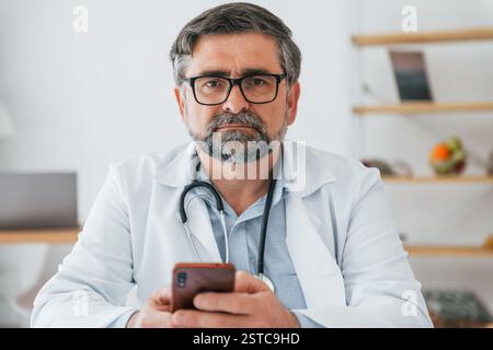 Le médecin masculin est assis avec le téléphone dans les mains. Un travailleur médical professionnel en manteau blanc est dans le bureau. Banque D'Images