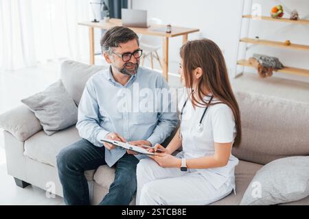 Médecin féminin est assis avec le client mature sur le canapé. Un travailleur médical professionnel en manteau blanc est dans le bureau. Banque D'Images