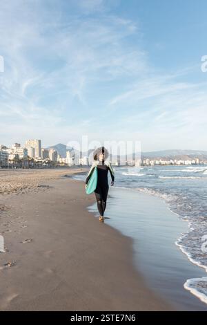 Jeune femme de surf attentionnés en combinaison avec planche de surf marchant en regardant loin sur le bord de mer lavé par la mer Banque D'Images