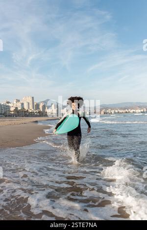 Jeune femme de surf attentionnés en combinaison avec planche de surf qui s'étourne sur la mer lavée par la mer Banque D'Images