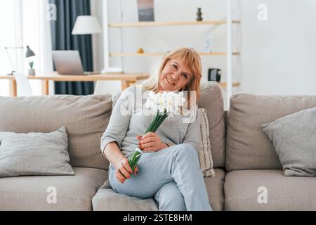 Assis sur un canapé avec des fleurs dans les mains. La femme âgée avec les cheveux roux est à la maison seule. Banque D'Images