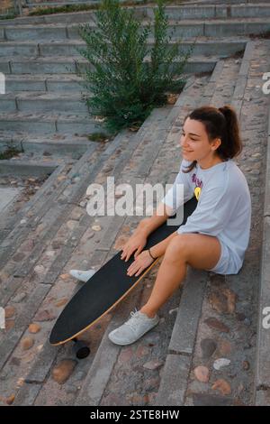 Une jeune femme détendue s'assoit sur les marches avec son skateboard lors d'une soirée urbaine tranquille Banque D'Images