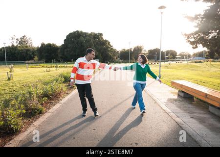 Un jeune couple latin se tenant joyeusement la main et marchant dans un parc, avec de longues ombres et la lumière du soleil filtrant à travers les arbres Banque D'Images