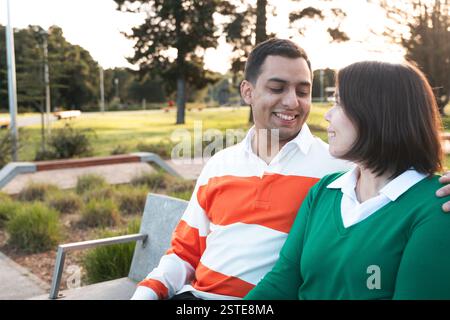 Un jeune couple latino se souriant, assis dehors dans un parc au crépuscule, reflétant une relation chaleureuse et aimante Banque D'Images