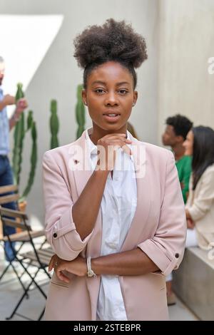 Femme afro-américaine confiante dans une tenue tendance debout sur la terrasse sur fond de groupe d'amis multiethniques et regardant la caméra Banque D'Images