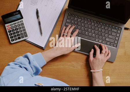 Mains vues taper sur un ordinateur portable avec un bloc-notes, un stylet et une calculatrice à proximité, démontrant l'efficacité du multitâche et du lieu de travail sur un bureau en bois Banque D'Images