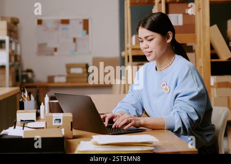 Femme hispanique travaillant sur ordinateur portable dans un espace de bureau moderne avec des étagères remplies de boîtes et de fournitures, concentrée sur sa tâche et utilisant des gadgets technologiques Banque D'Images