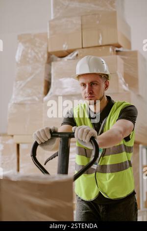 Ouvrier opérant un chariot élévateur dans un entrepôt, empilant des boîtes emballées sur des palettes en bois portant un gilet haute visibilité et un casque de sécurité Banque D'Images