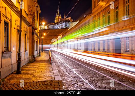 La rue Letenska brille la nuit tandis que les traînées de lumière des tramways créent une vue dynamique. Le château de Prague se profile en arrière-plan, mettant en valeur la beauté de la petite ville. Banque D'Images