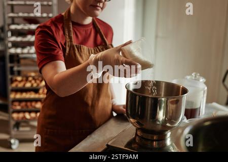 Plan recadré de boulangerie femelle versant des flocons de noix de coco de la tasse à mesurer dans le bol en acier inoxydable écaillant les ingrédients pour la pâte à pain dans la cuisine de la boulangerie, se concentrer sur les mains dans des gants en vinyle, copier l'espace Banque D'Images