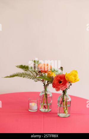 Des fleurs aux couleurs vives dans des vases en verre sont magnifiquement disposées sur une nappe rose, créant une atmosphère joyeuse. Banque D'Images