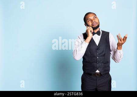 Chasseur afro-américain effectuant des réservations de chambres par appel téléphonique, partageant des informations sur les équipements de l'hôtel sur fond bleu. Le jeune portier répond aux questions en studio. Banque D'Images