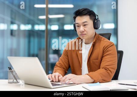 Sérieux homme asiatique confiant et concentré travaillant au bureau avec un ordinateur portable, portant des écouteurs, développeur programmeur masculin sur le lieu de travail, codant de nouveaux logiciels. Banque D'Images