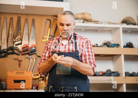 Un menuisier senior en chemise à carreaux rouge sable soigneusement un morceau de bois dans son atelier. Les outils pendent Banque D'Images