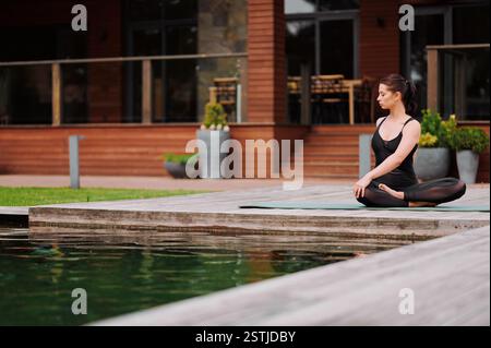 Jeune femme caucasienne effectue une torsion latérale assise dans la pose de lotus sur un tapis de yoga assis au bord de l'eau. Femme sportive pratiquant le yoga dans le parc. Femme doin Banque D'Images