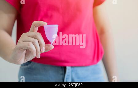 Femme portant une chemise rouge tenant une coupe menstruelle rose devant un fond blanc. Mise au point sélective. Banque D'Images