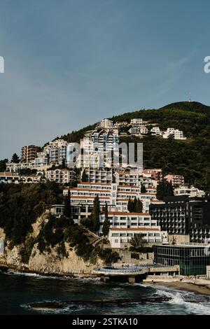 Ville côtière de Budva à flanc de colline avec des bâtiments modernes en terrasses surplombant la mer. Une végétation luxuriante entoure l'architecture, tandis que les vagues s'écrasent contre la Banque D'Images