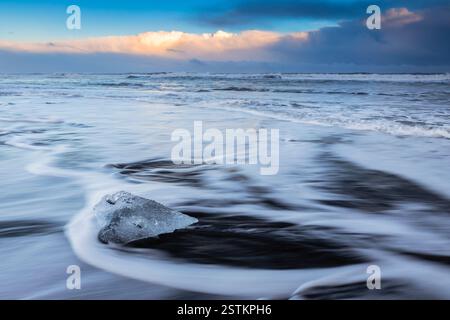 Vue sur les vagues de la mer qui coulent autour des blocs de glace au coucher du soleil sur la célèbre plage Diamond. Jökulsárlón, Diamond Beach, Austurland, Islande, Europe du Nord. Banque D'Images