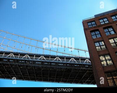 Une prise de vue en bas angle capture la structure métallique complexe du pont de Manhattan contre un ciel bleu clair. Adjacent au pont, une brique rouge classique Banque D'Images