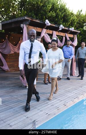 Amis seniors divers dansant joyeusement au bord de la piscine lors d'un événement de célébration dans le jardin. amitié, rassemblement, loisirs, bonheur Banque D'Images