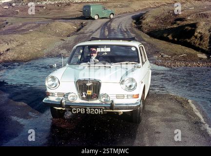 Pilote femme non identifiée dans les années 1970 conduisant à travers une petite rivière dans une zone déserte avec une camionnette verte en arrière-plan, dans une voiture vintage blanche avec des badges de club de voiture sur la grille du radiateur avant. Photographie des années soixante-dix. Banque D'Images