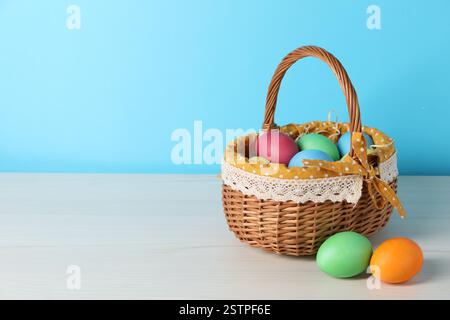 Beaux oeufs de Pâques décorés dans un panier en osier sur une table en bois blanc sur fond bleu clair, espace pour le texte Banque D'Images