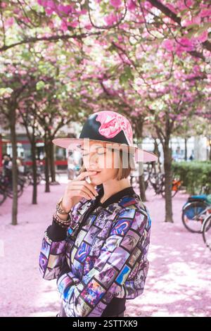 Belle femme portant une tenue lila à la mode et un chapeau peint debout dans l'allée des fleurs de cerisier en Allemagne Cologne. Banque D'Images