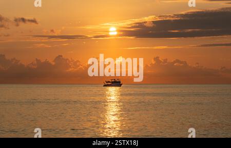 Un bateau glisse sur des eaux tranquilles au coucher du soleil, avec la lumière dorée du soleil qui se reflète sur la mer. Des nuages spectaculaires encadrent le ciel Banque D'Images