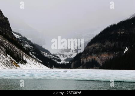 Un paysage spectaculaire avec un lac partiellement gelé entouré de montagnes imposantes et enveloppé de brume, créant une atmosphère mystérieuse Banque D'Images