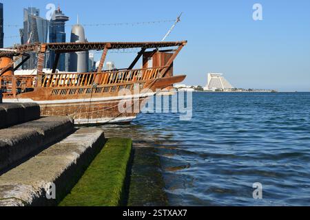 Bateau à boutres traditionnel en bois amarré le long de la corniche de Doha avec l'horizon futuriste de la ville, créant un contraste saisissant entre le bateau maritime du Qatar Banque D'Images