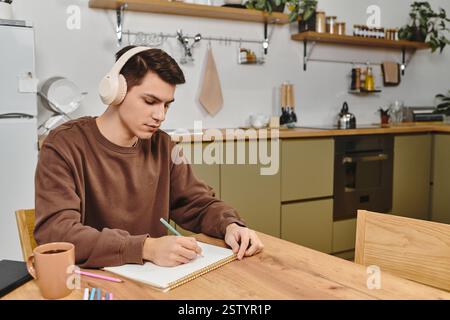 Un jeune homme dessine à une table en bois dans une cuisine confortable, écouteurs. Banque D'Images
