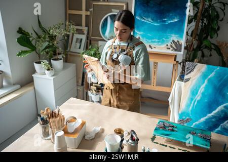 Artiste peintre féminin focalisé prépare un panneau de bois pour l'application de résine époxy dans un atelier d'art. Banque D'Images