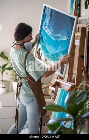 L'artiste créatrice réfléchie place la peinture époxy finie sur chevalet en bois dans l'atelier d'art. Banque D'Images