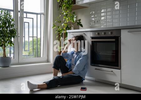 Malheureuse fille asiatique assise sur le sol dans la cuisine se sentant déprimé après l'utilisation des médias sociaux Banque D'Images