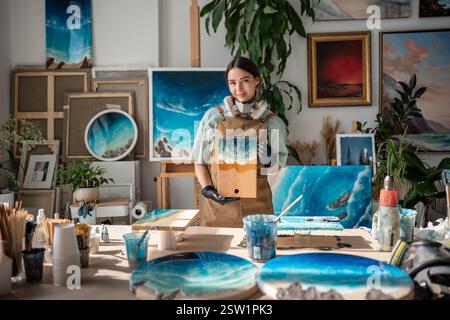 Heureuse femme satisfaite artiste tient la peinture de mer de résine époxy regarde les stands d'appareil photo dans le studio d'art. Banque D'Images
