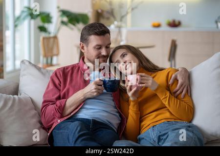 Heureux couple serrant sur le canapé, buvant du café, du thé. Conjoints satisfaits se relaxant dans une étreinte chaleureuse. Banque D'Images