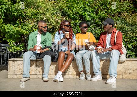 Groupe détendu d'amis afro-américains assis sur le banc du parc buvant du café tout en discutant Banque D'Images